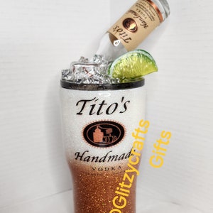 May include: A white and copper glitter tumbler with a Tito's Handmade Vodka logo and the text "@Glitzycrafts Gifts". A bottle of Tito's Vodka is upside down in the tumbler with a lime wedge and ice.