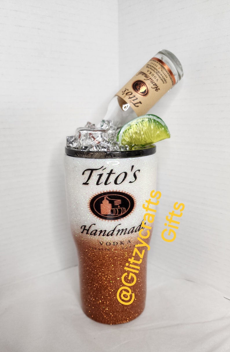 May include: A white and copper glitter tumbler with a Tito's Handmade Vodka logo and the text "@Glitzycrafts Gifts". A bottle of Tito's Vodka is upside down in the tumbler with a lime wedge and ice.