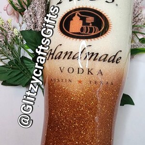 May include: A white and gold glitter tumbler with a Tito's Handmade Vodka logo. The logo features a black and orange illustration of a still and barrels. The text "Tito's Handmade Vodka" is printed in black on the tumbler. The text "Austin, Texas" is printed in black at the bottom of the tumbler.