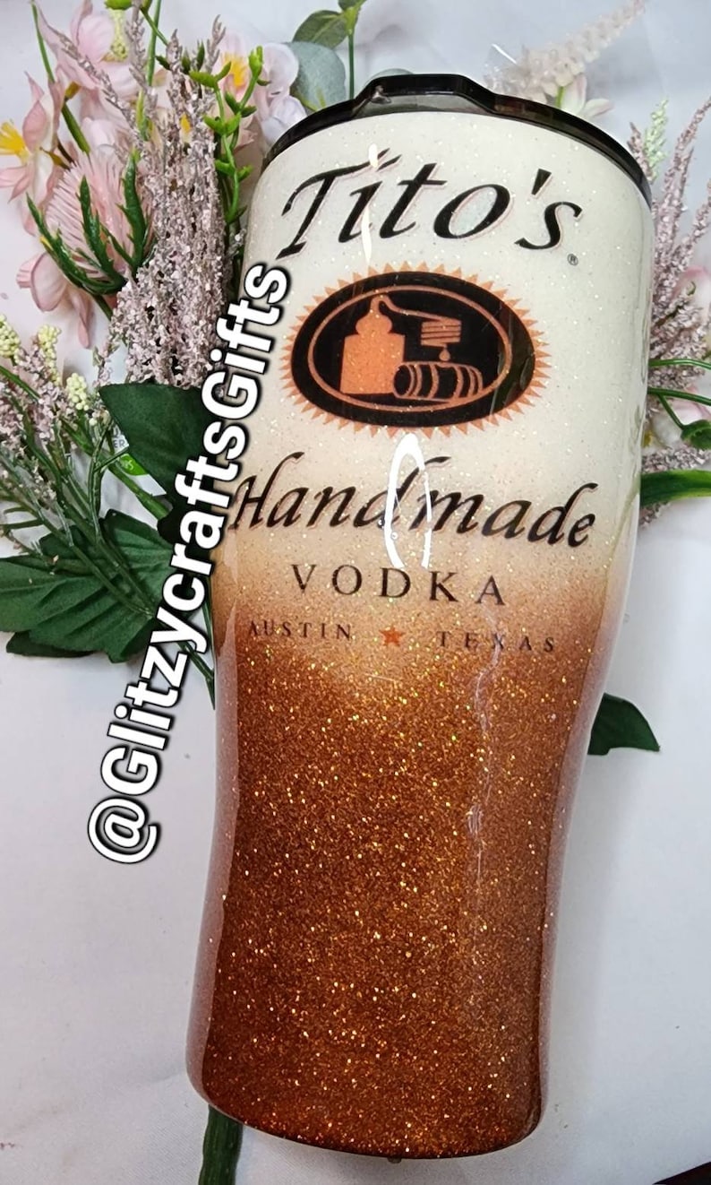 May include: A white and gold glitter tumbler with a Tito's Handmade Vodka logo. The logo features a black and orange illustration of a still and barrels. The text "Tito's Handmade Vodka" is printed in black on the tumbler. The text "Austin, Texas" is printed in black at the bottom of the tumbler.