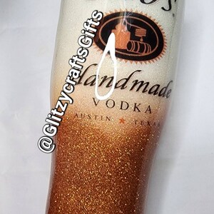May include: A white and gold glitter tumbler with a Tito's Handmade Vodka logo. The logo features a black and white illustration of a still with barrels and the text "Tito's Handmade Vodka Austin Texas".