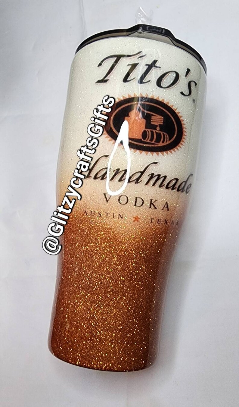 May include: A white and gold glitter tumbler with a Tito's Handmade Vodka logo. The logo features a black and white illustration of a still with barrels and the text "Tito's Handmade Vodka Austin Texas".