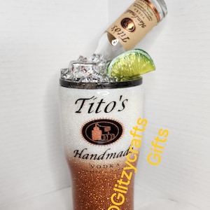 May include: A white and copper glitter tumbler with a Tito's Handmade Vodka logo and the text "@Glitzycrafts Gifts". A bottle of Tito's Vodka is upside down in the tumbler with a lime wedge and ice.