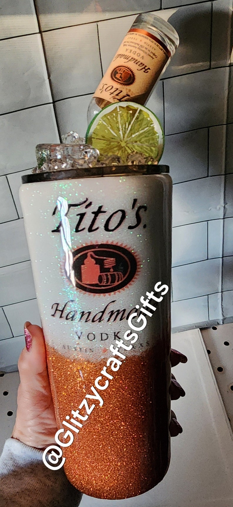 May include: A white and orange glitter tumbler with the text "Tito's Handmade Vodka" and the logo of Tito's Vodka. The tumbler is topped with ice and a lime wedge. The tumbler is held by a hand with pink nail polish. The text "@GlitzycraftsGifts" is on the side of the tumbler.