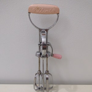May include: A vintage hand-crank mixer with a pink plastic handle and the word "Flint" printed on it. The mixer has three metal beaters and a chrome finish.