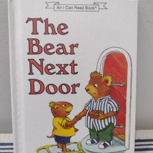 May include: A children's book cover with the title "The Bear Next Door" by Ida Luttrell and illustrations by Sarah Stapler. The cover features a brown bear holding a plate of cookies and a smaller brown bear with yellow shirt.