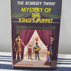 May include: A hardcover book titled The Bobbsey Twins' Mystery of the King's Puppet by Laura Lee Hope. The cover features a dark purple stage with curtains, a knight puppet, and four children standing in front of the stage in a theater setting.