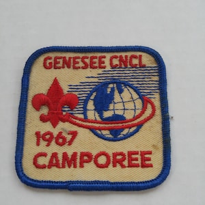 May include: A vintage patch with a tan background and a blue border. The patch features the text "GENESEE CNCL" and "1967 CAMPOREE" in red. A red fleur-de-lis and a blue globe with a red orbit are also present.