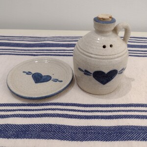 May include: A white ceramic pitcher with a blue heart design and a cork stopper. The pitcher is sitting on a white and blue striped surface.