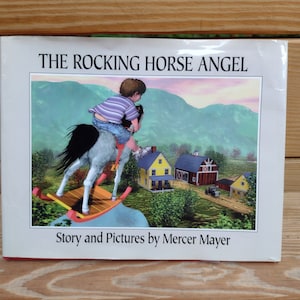 May include: A book titled "The Rocking Horse Angel" with a colorful illustration of a child riding a rocking horse in a rural setting. The text "Story and Pictures by Mercer Mayer" is printed below the illustration.