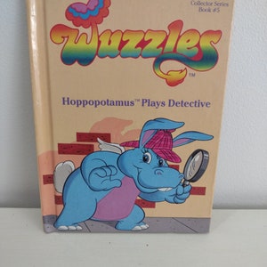 May include: A book cover with a rainbow colored word "Wuggles" and a blue hippopotamus wearing a red hat and holding a magnifying glass. The book is titled "Hippopotamus Plays Detective" and is part of the "Collector Series" book #5.