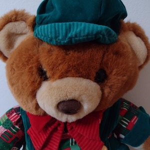 May include: A brown teddy bear wearing a green and red plaid shirt, a red bow tie, and a green velvet hat. The bear is a Christmas-themed plush toy.