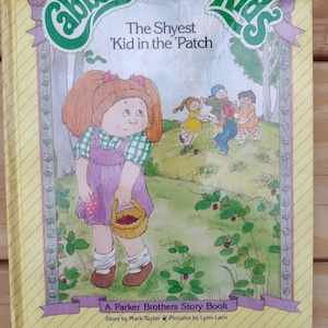 May include: A book cover featuring a Cabbage Patch Kid doll with red hair and a purple dress. The title of the book is "The Shyest 'Kid in the 'Patch." The book is a Parker Brothers Story Book, written by Mark Taylor and illustrated by Lynn Lace.