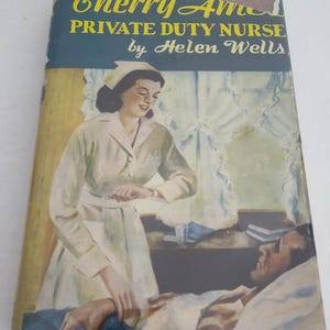 May include: A vintage book cover featuring a nurse in a white uniform and a white cap, looking down at a man lying in bed. The book title is "Cherry Ames, Private Duty Nurse" by Helen Wells.