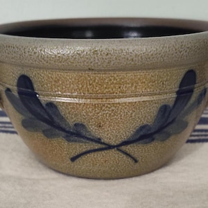 May include: A stoneware bowl with a speckled tan exterior and a dark brown interior. The bowl features two handles and is decorated with blue leaf designs. The rim has a textured, light brown band.