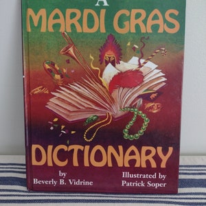 May include: A book titled "A Mardi Gras Dictionary" by Beverly B. Vidrine and illustrated by Patrick Soper. The cover features a colorful illustration of a Mardi Gras mask, beads, and other festive items.