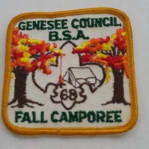 May include: A white embroidered patch with a green border and a yellow outer border. The patch features a scout's symbol with a tent and a campfire. The text "Genesee Council B.S.A. Fall Camporee" is embroidered on the patch.