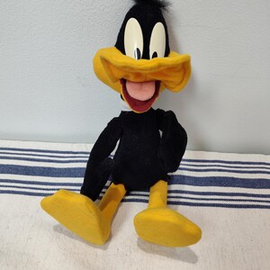 May include: A plush Daffy Duck toy, featuring a black body, yellow beak and feet, and large white eyes. The cartoon character has a red tongue sticking out and is sitting on a striped surface.