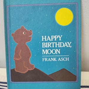 May include: A children's book titled "Happy Birthday, Moon" by Frank Asch. The cover features a brown bear looking up at a yellow moon against a blue starry sky. The book has a teal border.