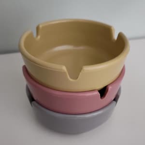 May include: A stack of three ceramic ashtrays in pastel colors. The top ashtray is a light yellow, the middle one is pink, and the bottom one is gray. Each ashtray has a curved design with a notch for resting cigarettes.