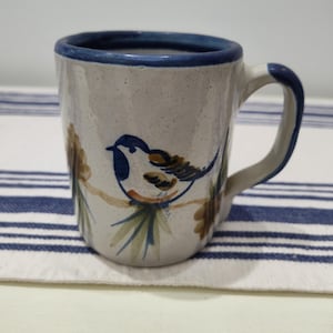 May include: A speckled off-white ceramic mug featuring a hand-painted blue and brown bird perched on a branch. The mug has a thick blue rim and a matching blue handle. The exterior surface displays a subtle speckled texture throughout the clay body.