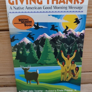 May include: A children's book cover with a Native American theme. The cover features a colorful illustration of a mountain landscape with a deer, a river, and a group of children. The title of the book is "Giving Thanks: A Native American Good Morning Message".