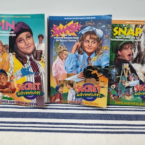 May include: Three vintage paperback books from the "Secret Adventures" series. The book covers feature illustrations of young people and text including "Spin", "Smash", and "Snap". The books are on a blue and white striped surface.