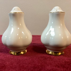 May include: A pair of white ceramic salt and pepper shakers with gold trim. The shakers are shaped like a bulb with a rounded top and a flared base.