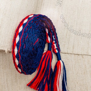 May include: A blue, red, and white braided cord with tassels. The cord is woven with a diamond pattern. The cord is on a white linen background. The text "Medieval Armor & Craft www.lembergarmor.com" is visible in the background.
