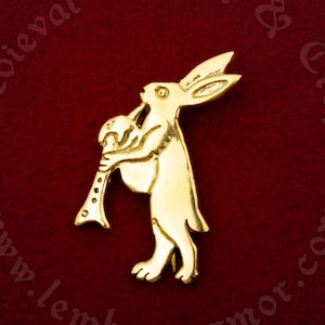 Puede incluir: Un pin de metal dorado que representa un conejo tocando un instrumento musical. El conejo está de pie sobre sus patas traseras y sostiene una flauta en sus patas delanteras. El pin está sobre un fondo de terciopelo rojo.