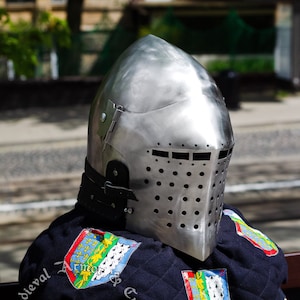 May include: A silver helmet with a visor and a black leather strap. The helmet is sitting on a black fabric with colourful patches that say "Medieval Armor & Craft" and "facebook: Lembergshop".