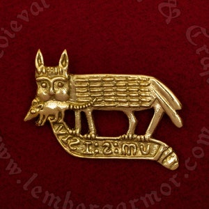 Könnte beinhalten: Eine goldfarbene Metallnadel, die einen Wolf darstellt, der ein kleineres Tier im Maul trägt. Der Wolf steht auf einem gebogenen Sockel mit der Inschrift "Ave Maria" in lateinischer Sprache.