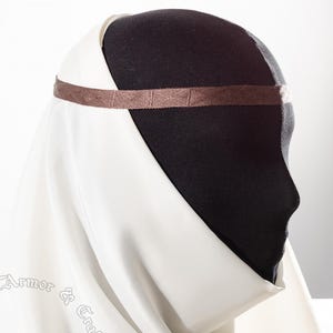 May include: A white medieval veil and head covering, draped over a black mannequin head. A brown band is wrapped around the head. The veil is made of a soft, flowing fabric. The image is set against a white background.