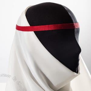 May include: A black mannequin head is draped with a white fabric and a red headband. The fabric is draped over the head and shoulders. The headband is a thin, textured band that sits across the forehead. The overall look is reminiscent of medieval attire.