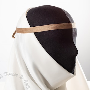 May include: A white medieval coif with a tan headband and trim. The coif is draped over a black mannequin head. The headband is secured with a small braided cord. The coif is made of a soft, flowing fabric.