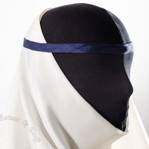 May include: A white medieval coif with a dark blue headband, displayed on a black mannequin head. The coif is made of a flowing fabric, and the headband is a simple band.