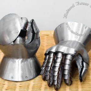 Könnte beinhalten: Ein Paar mittelalterliche Handschuhe aus Metall. Die Handschuhe sind silberfarben und haben eine strukturierte Oberfläche. Die Handschuhe sind so konzipiert, dass sie die Hände und Finger im Kampf schützen.