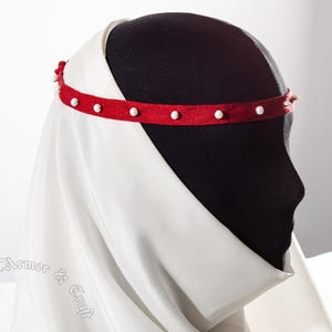 May include: A red medieval headband with white pearl-like beads. The headband is worn over a white veil and is displayed on a black mannequin head. The image is a side profile, showcasing the historical design.