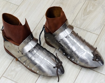 May include: A pair of brown leather boots with metal armour plating. The boots have laces and are pointed at the toe. The boots are on a light brown tiled surface. The text "Medieval Armor & Craft" is visible in the background.