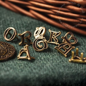 Monturas medievales de oro (latón) con letras latinas. Adornos y alfileres con el alfabeto para cinturón y traje medieval.