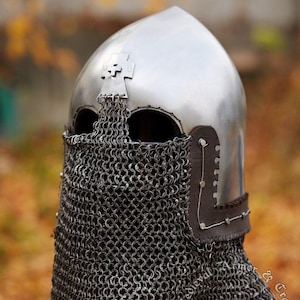 May include: A silver helmet with a chainmail face mask. The helmet has a pointed top and a cross on the front. The chainmail mask covers the face and neck. The helmet is on a wooden stand. www.medievalarmour.eu