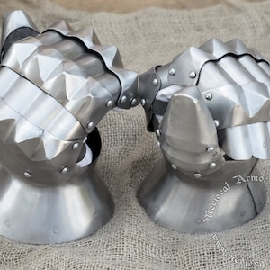 Könnte beinhalten: Ein Paar mittelalterliche Handschuhe mit Stacheln an den Knöcheln. Die Handschuhe sind aus Metall und haben eine silberne Oberfläche. Die Handschuhe liegen auf einem braunen Jutesack-Hintergrund.