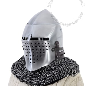 Puede incluir: Un casco plateado con una parte superior puntiaguda y una máscara facial con un diseño de malla. El casco se lleva sobre un collar de cota de malla.