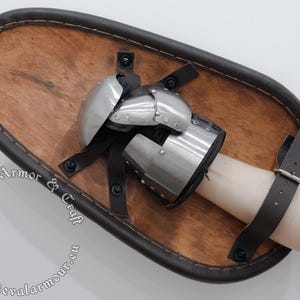 May include: A medieval-style shield with a wooden core and a brown leather border. The shield features a metal gauntlet attached to the center, secured with leather straps. The shield is designed for historical reenactment or display.