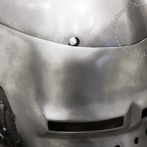 May include: A close-up of a silver helmet with a visor and a small vent on the side. The helmet is made of metal and has a textured surface. The text "Medieval Armor & Craft" is visible on the helmet.