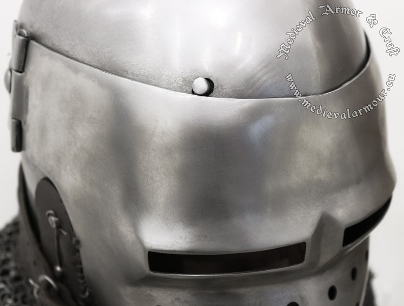 May include: A close-up of a silver helmet with a visor and a small vent on the side. The helmet is made of metal and has a textured surface. The text "Medieval Armor & Craft" is visible on the helmet.