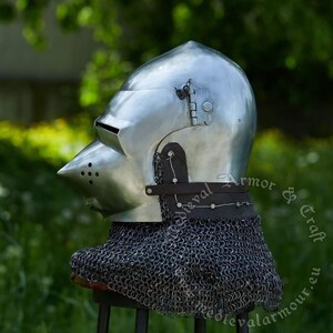 May include: A silver helmet with a pointed visor and a chainmail coif. The helmet has a hinged visor and a leather strap. The helmet is on a stand and is in front of a green background.