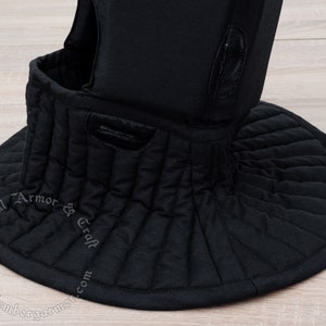 May include: Black padded medieval style neck guard with a quilted design. The neck guard is made of fabric and has a round shape. The text "Medieval Armor & Craft" is printed on the guard.
