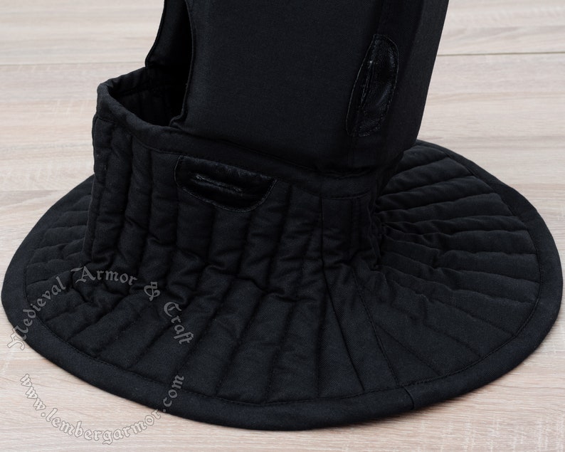 May include: Black padded medieval style neck guard with a quilted design. The neck guard is made of fabric and has a round shape. The text "Medieval Armor & Craft" is printed on the guard.