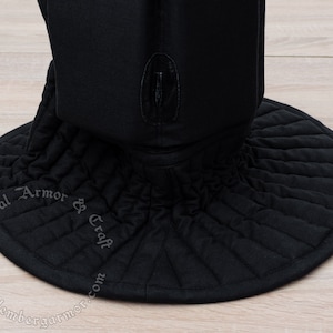 May include: Black quilted fabric medieval coif with a wide, round brim. The coif has a small, black leather tab on the top. The text "Medieval Armor & Craft" is embroidered on the brim.
